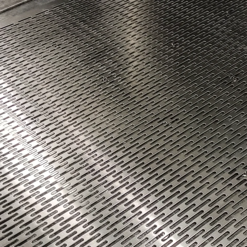 Zbliżenie na arkusz blachy stalowej poddanej precyzyjnej perforacji z&nbsp;gęstym wzorem podłużnych otworów.